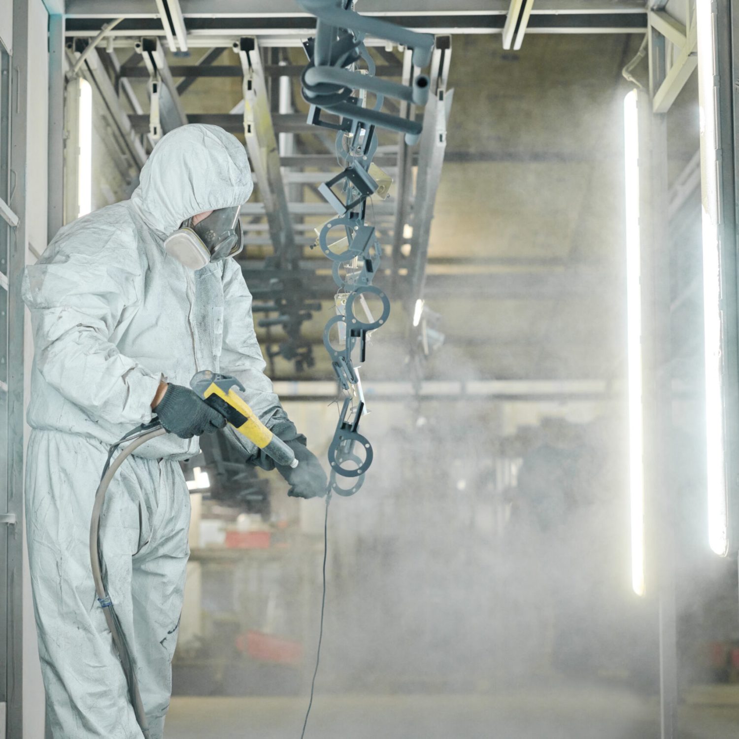 Man in protective suit spraying powder paint from gun on metal products at factory